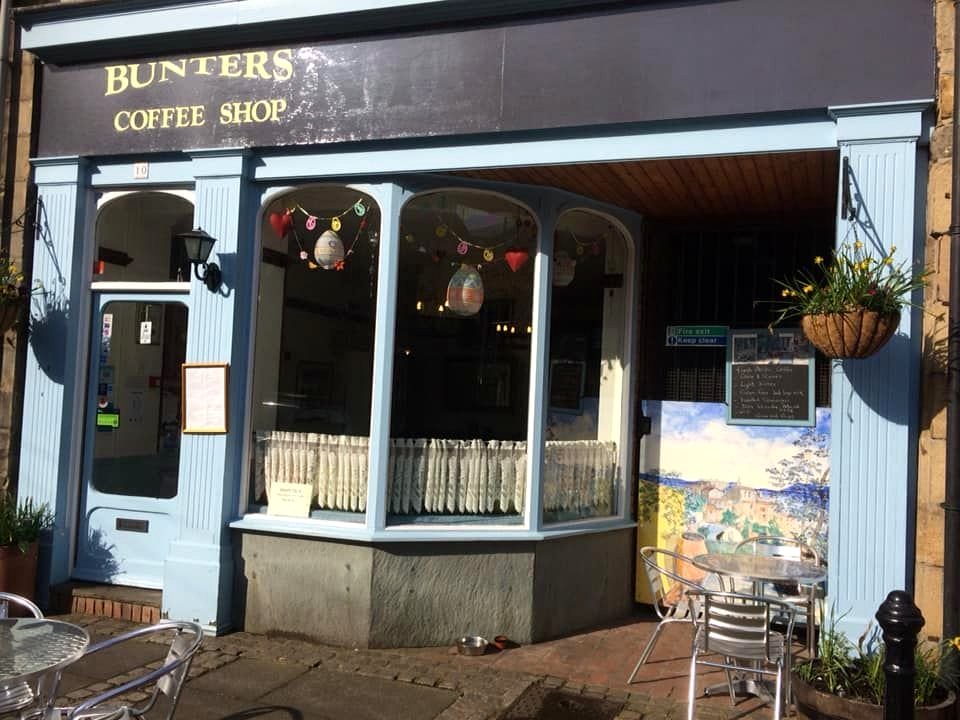 The bay window of Bunters, with lace half-curtains, bunting and hanging baskets, and chairs set out on the pavement