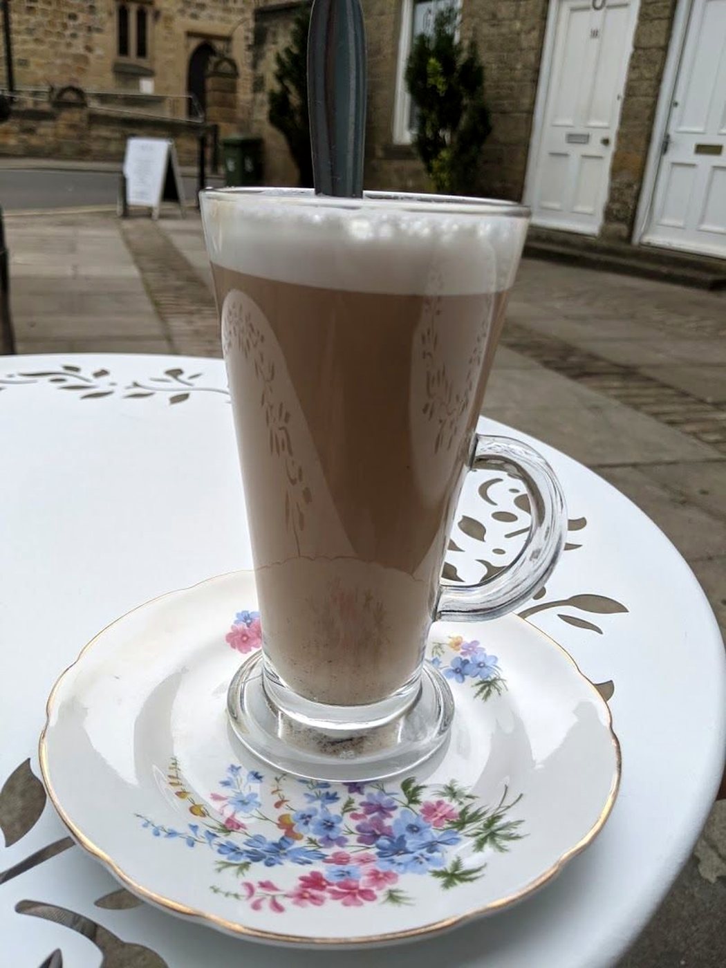 A latte in a tall glass on a white china saucer with blue and pink flowers, on a white table outside the café